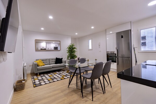 A modern kitchen with a built-in refrigerator, a dining area with a glass table and chairs, and a gray sofa with a yellow pillow. - Apartamento Moderno + Parking (Oviedo)