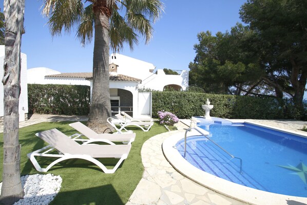 A swimming pool with lounge chairs, a palm tree, and a white building with a tiled roof.