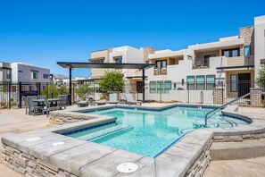 A modern outdoor pool area with lounge chairs and a hot tub, surrounded by a fenced patio with a view of residential buildings.
