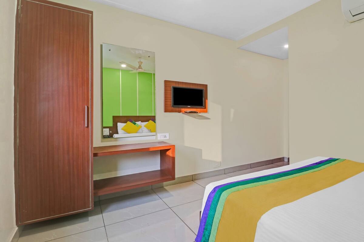 A hotel room with a wooden wardrobe, a small desk with a mirror, a television mounted on the wall, and a bed with a colorful bedspread.