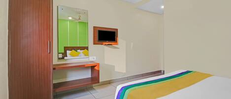 A hotel room with a wooden wardrobe, a small desk with a mirror, a television mounted on the wall, and a bed with a colorful bedspread.