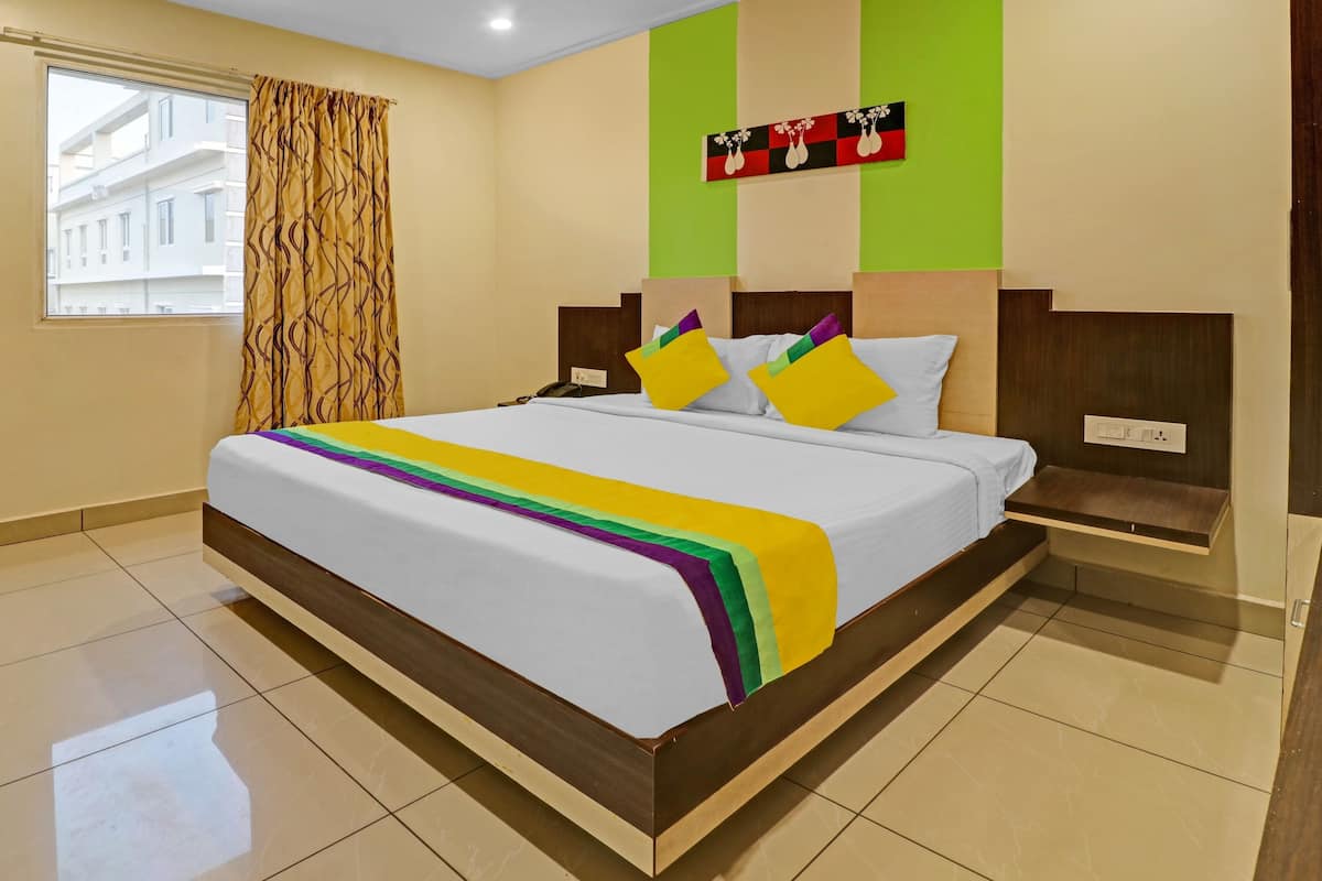 A hotel room with a large bed, a bedside table, a chair, a window with curtains, and a wall with a painting.