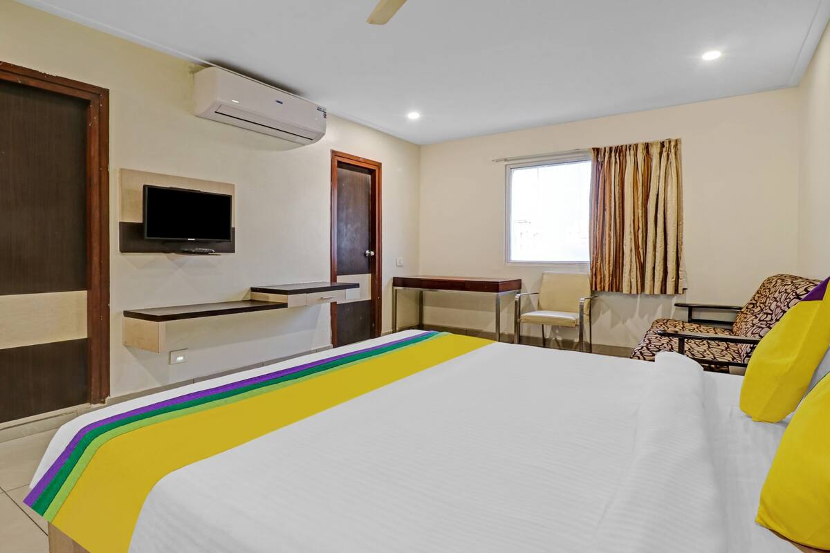 A hotel room with a bed, a TV mounted on the wall, a desk, a chair, and a window with curtains.