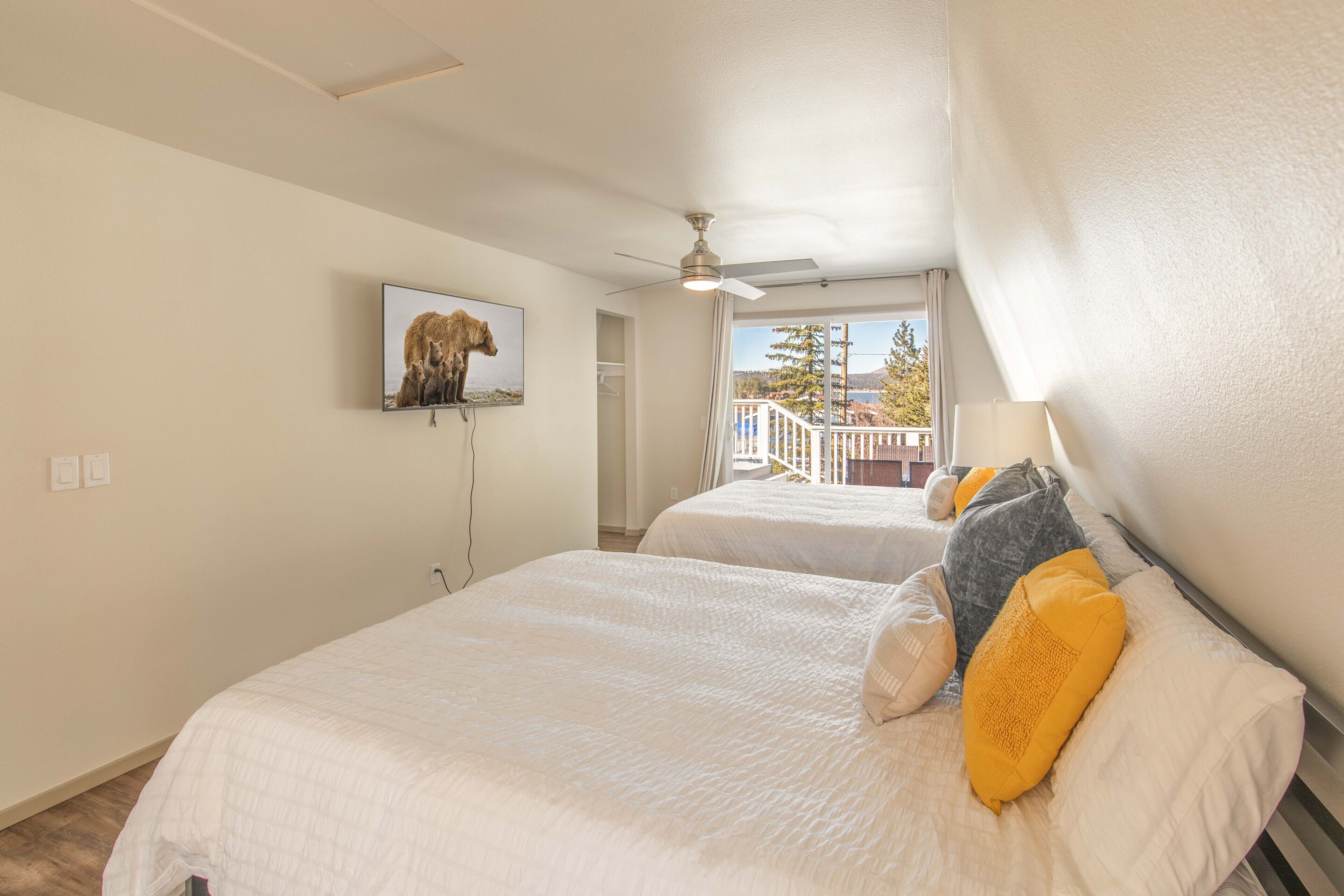 A bedroom with two beds, a ceiling fan, a mounted TV, and a balcony with a railing.