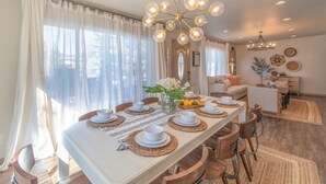 Comedor con una mesa larga dispuesta para cuatro, rodeada de sillas de madera. Un candelabro cuelga encima, y la habitación tiene ventanas grandes con cortinas de gasa.