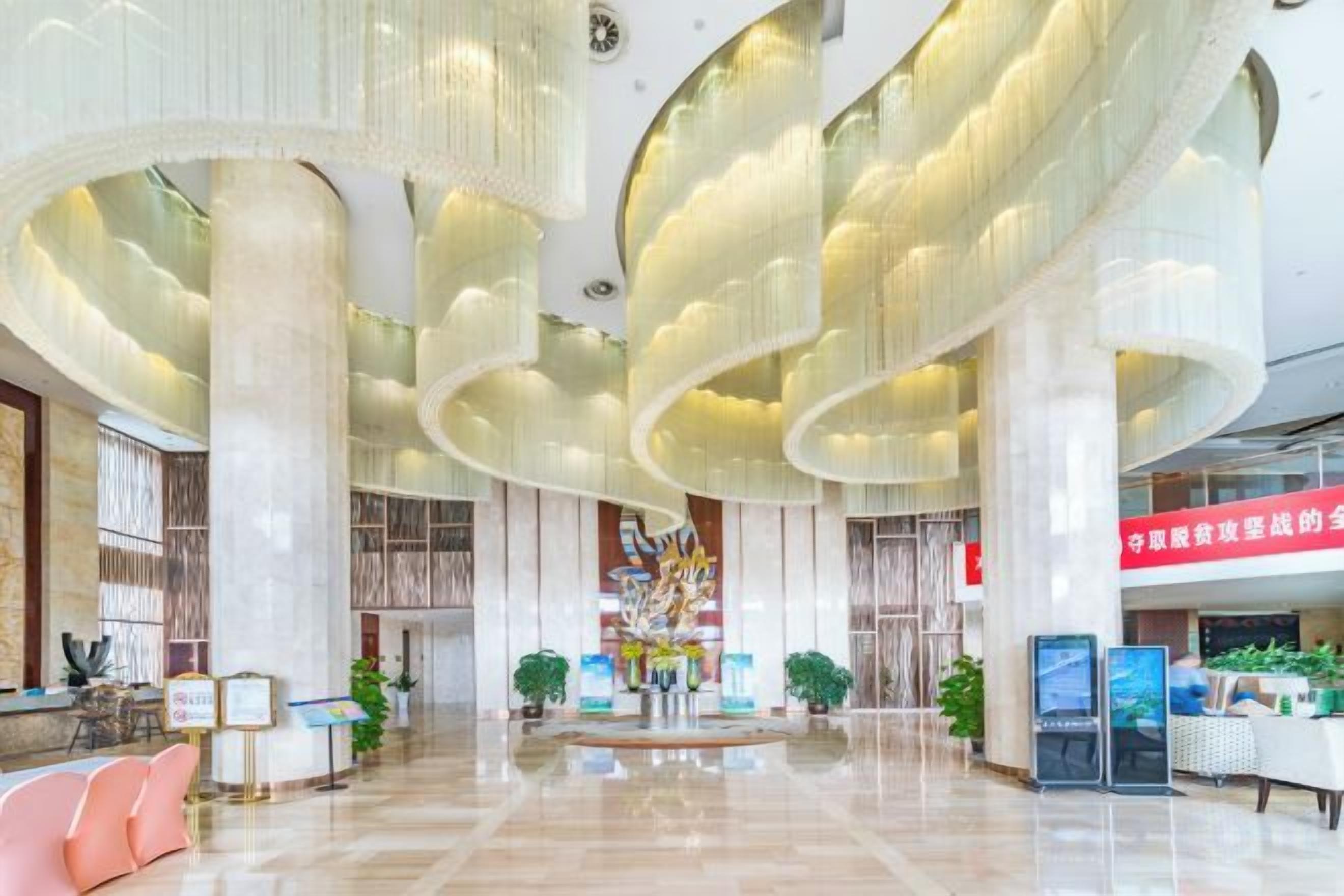 A spacious lobby with a central reception desk, large columns, and a modern chandelier.