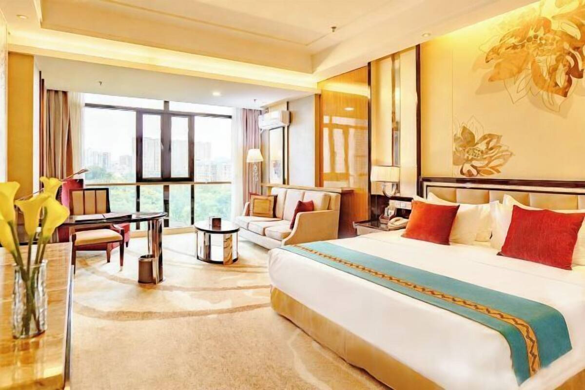 A hotel room with a large bed, a sofa, a desk, and a view of the city through large windows.