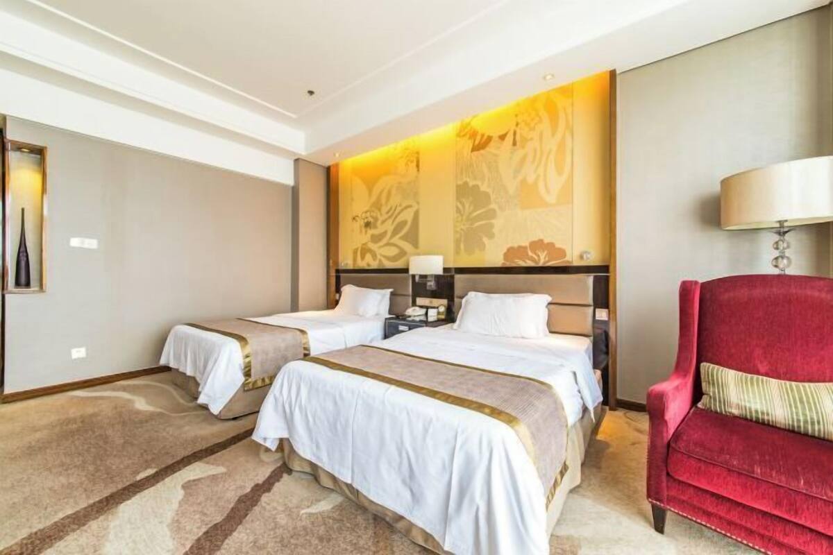 A hotel room with two beds, a red armchair, a large floral wall mural, and a lamp.