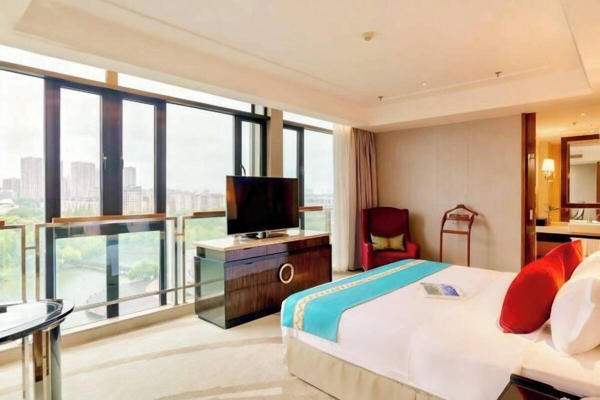 A hotel room with a large bed, a television on a wooden dresser, a chair, and a view of the city through large windows.