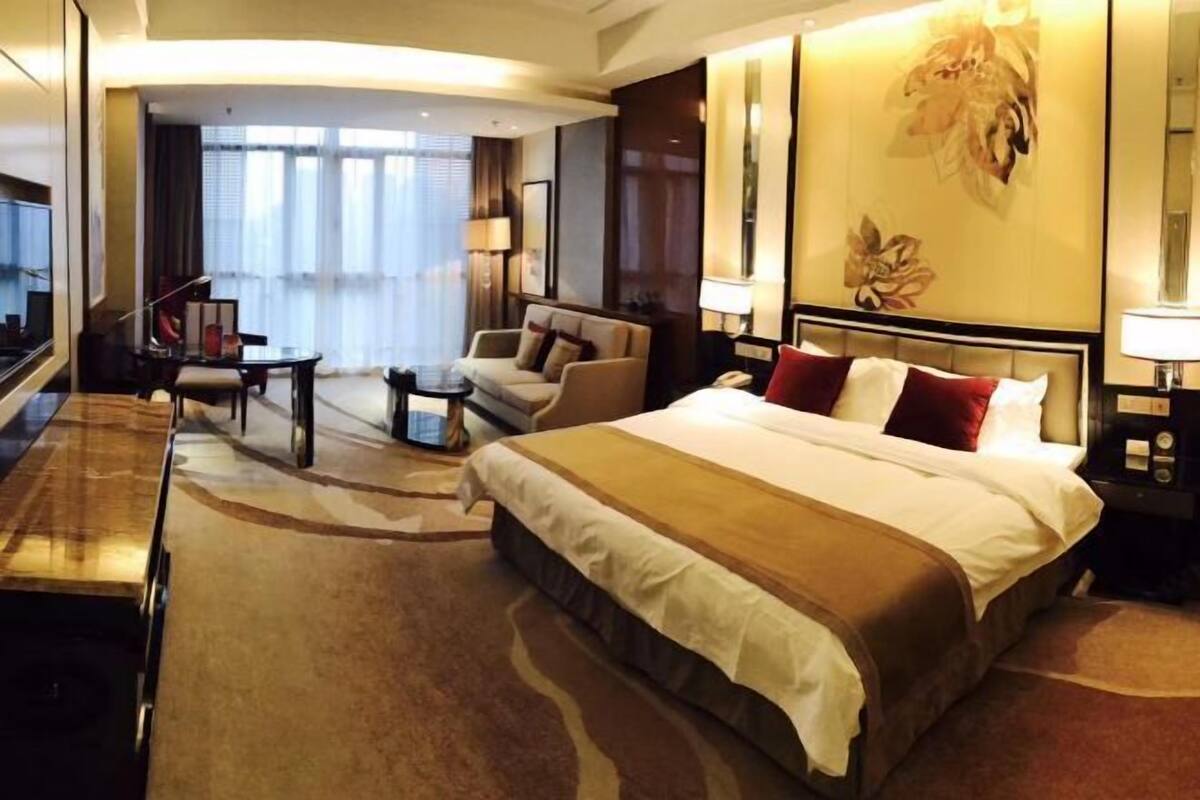 A hotel room with a large bed, a sitting area with a sofa and armchairs, a small table, and a large window with curtains.