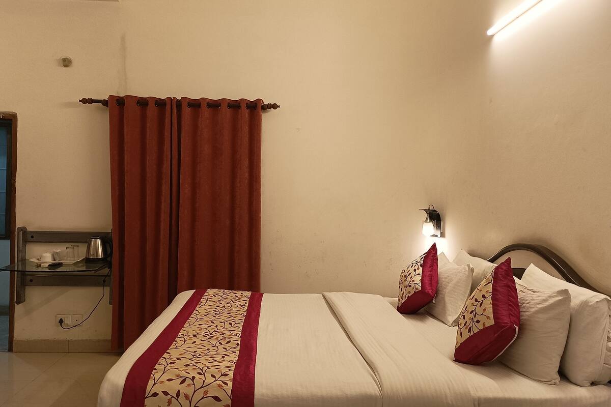Un llit ben fet amb un estovat estampat i almoines, una taula de nit amb una làmpada, i una finestra amb cortines.