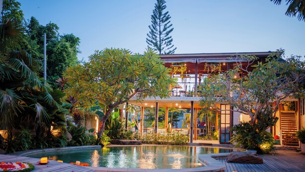 Um edifício moderno com piscina, cercado por vegetação exuberante e um deque de madeira.