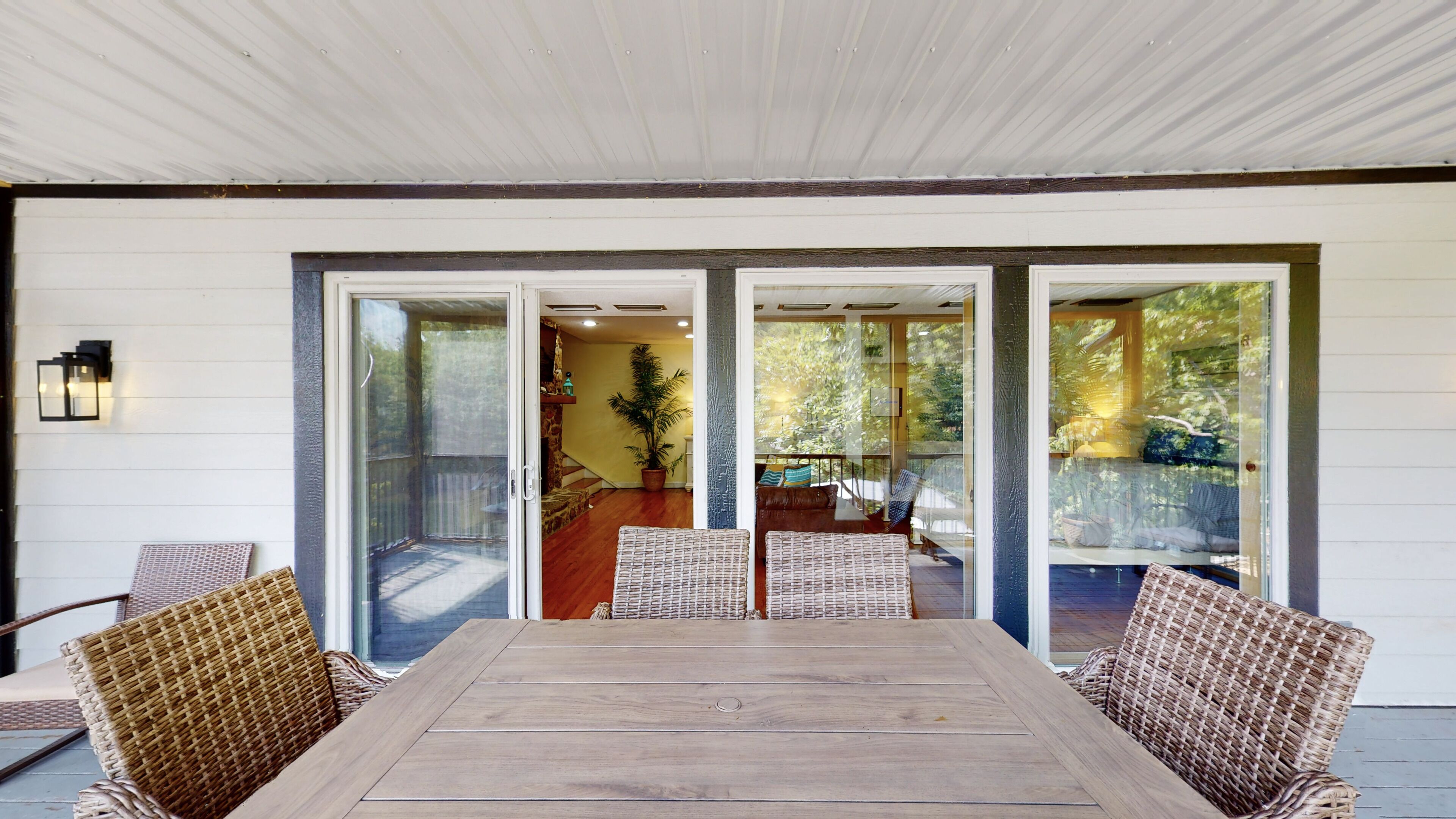 A covered patio with a wooden table and wicker chairs, glass sliding doors leading to an interior space with a potted plant and a view of trees.