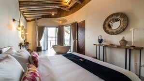 Ein geräumiges Schlafzimmer mit einem großen Bett, einer Holzbalkendecke, einem runden Spiegel und Blick ins Freie.