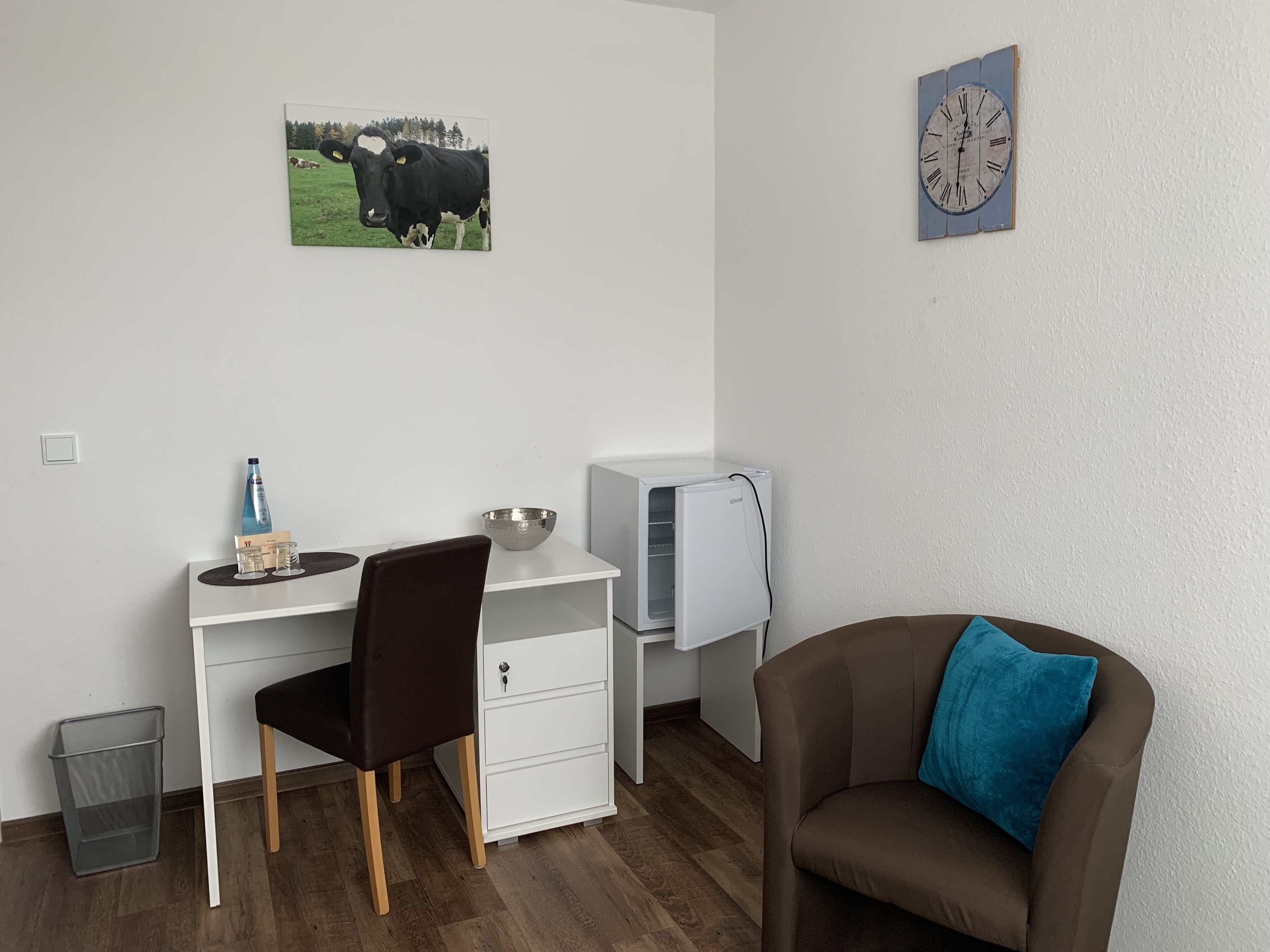 Ein kleiner, ordentlicher Raum mit Schreibtisch, Stuhl und einem braunen Sessel mit blauer Kissen. Ein kleiner Mülleimer, ein weißer Kühlschrank und eine Wanduhr sind ebenfalls vorhanden.