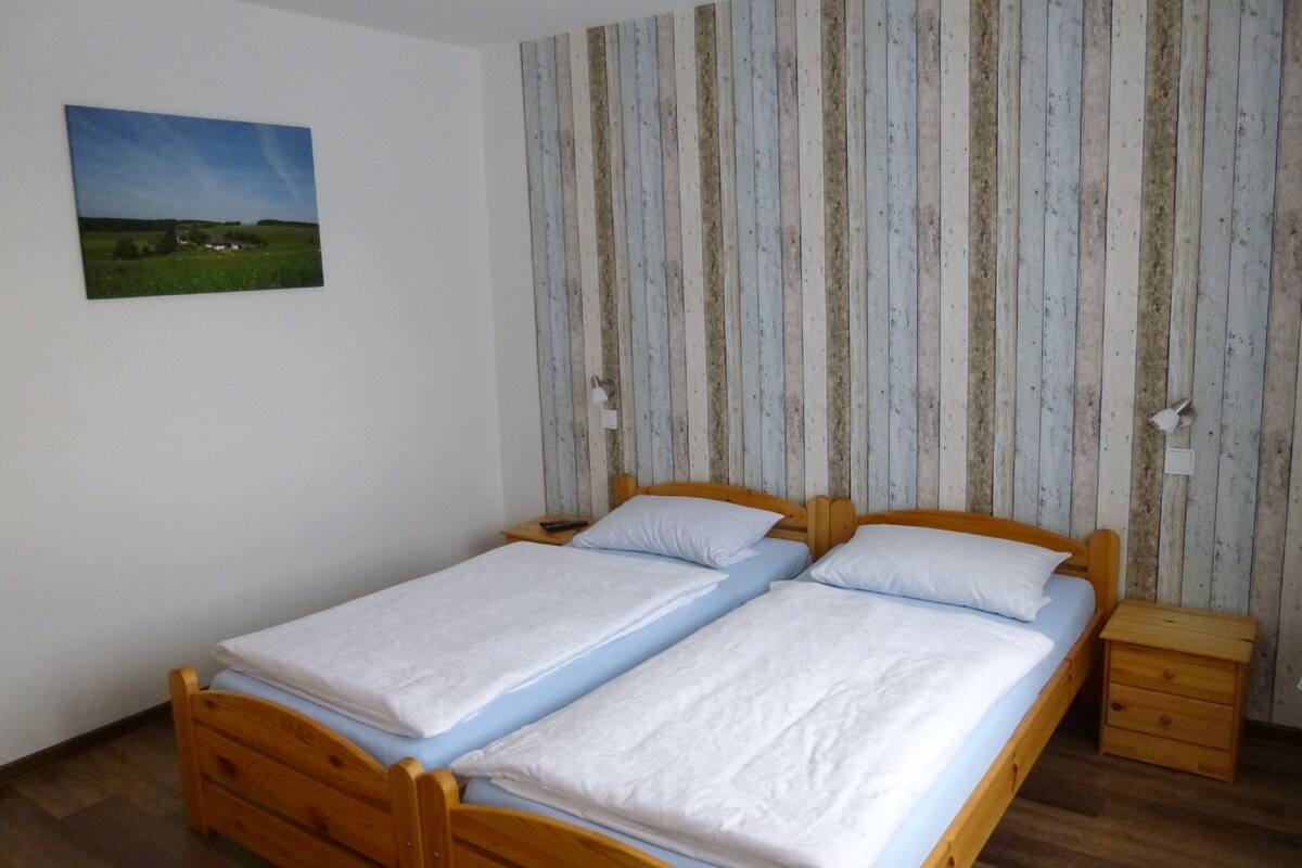 A room with two wooden beds, a small wooden nightstand, a wall-mounted painting, and a striped wallpaper background.