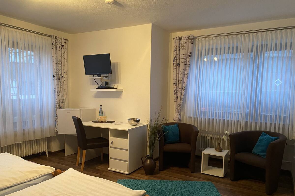 A hotel room with a bed, a desk with a computer, two chairs, a small table, and a window with sheer curtains.