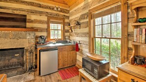 A rustic cabin interior with a stone fireplace, wooden walls, a kitchen with a sink and stove, and a large window overlooking greenery. - Numerous On-Site Trails: Earlysville Wooded Cabin (Earlysville)