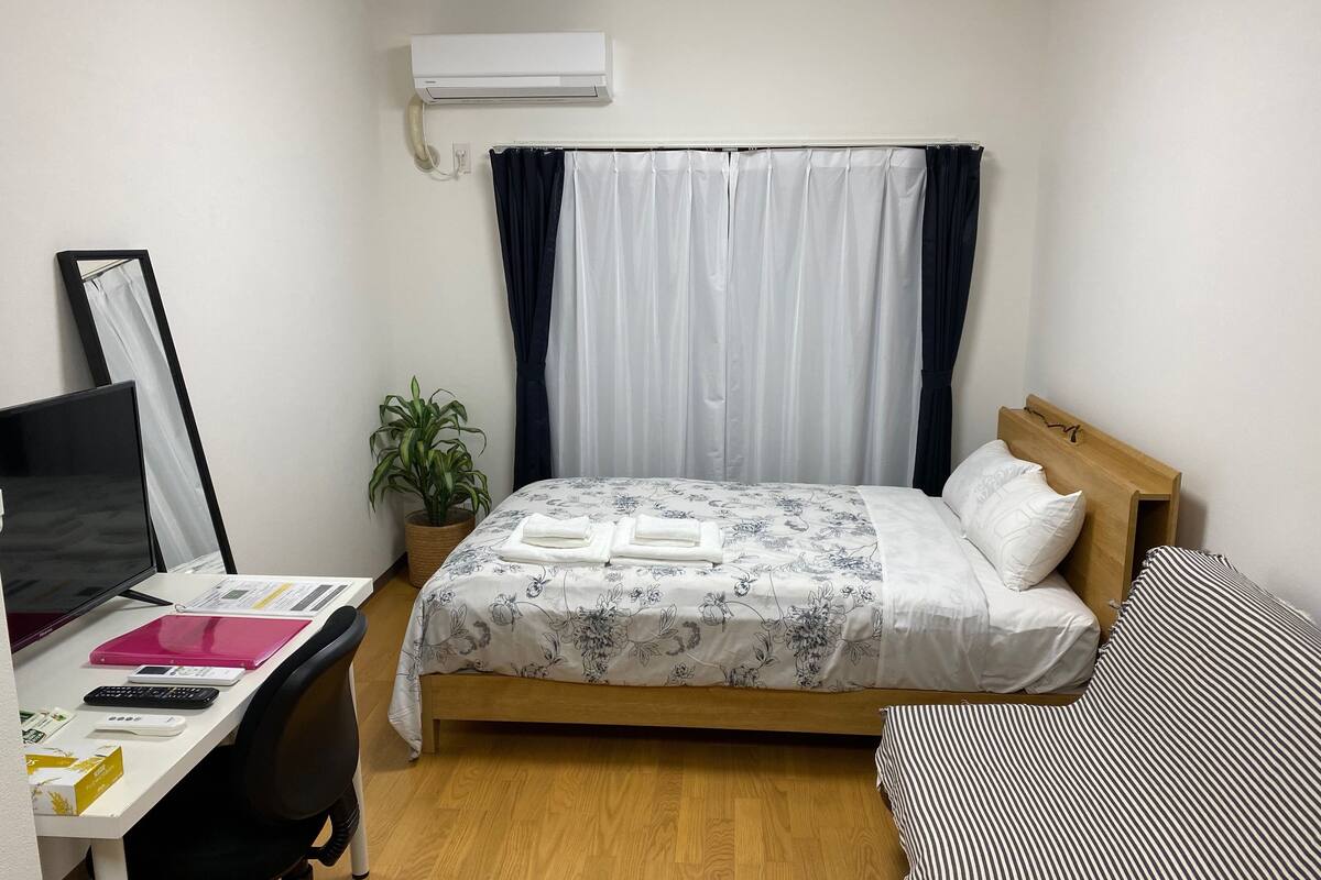 A bedroom with a bed, a desk with a computer, a potted plant, and a window with curtains.