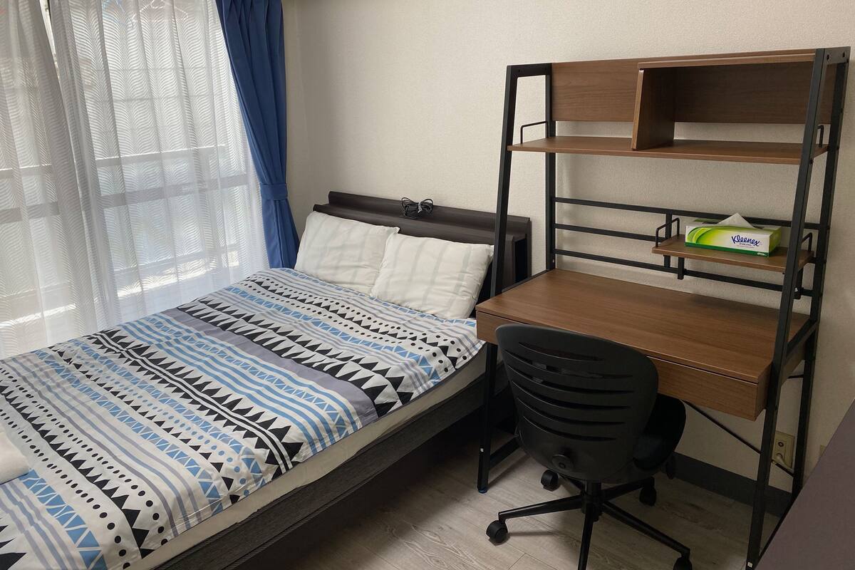 A bedroom with a bed, a desk with a chair, and a shelf with a tissue box.
