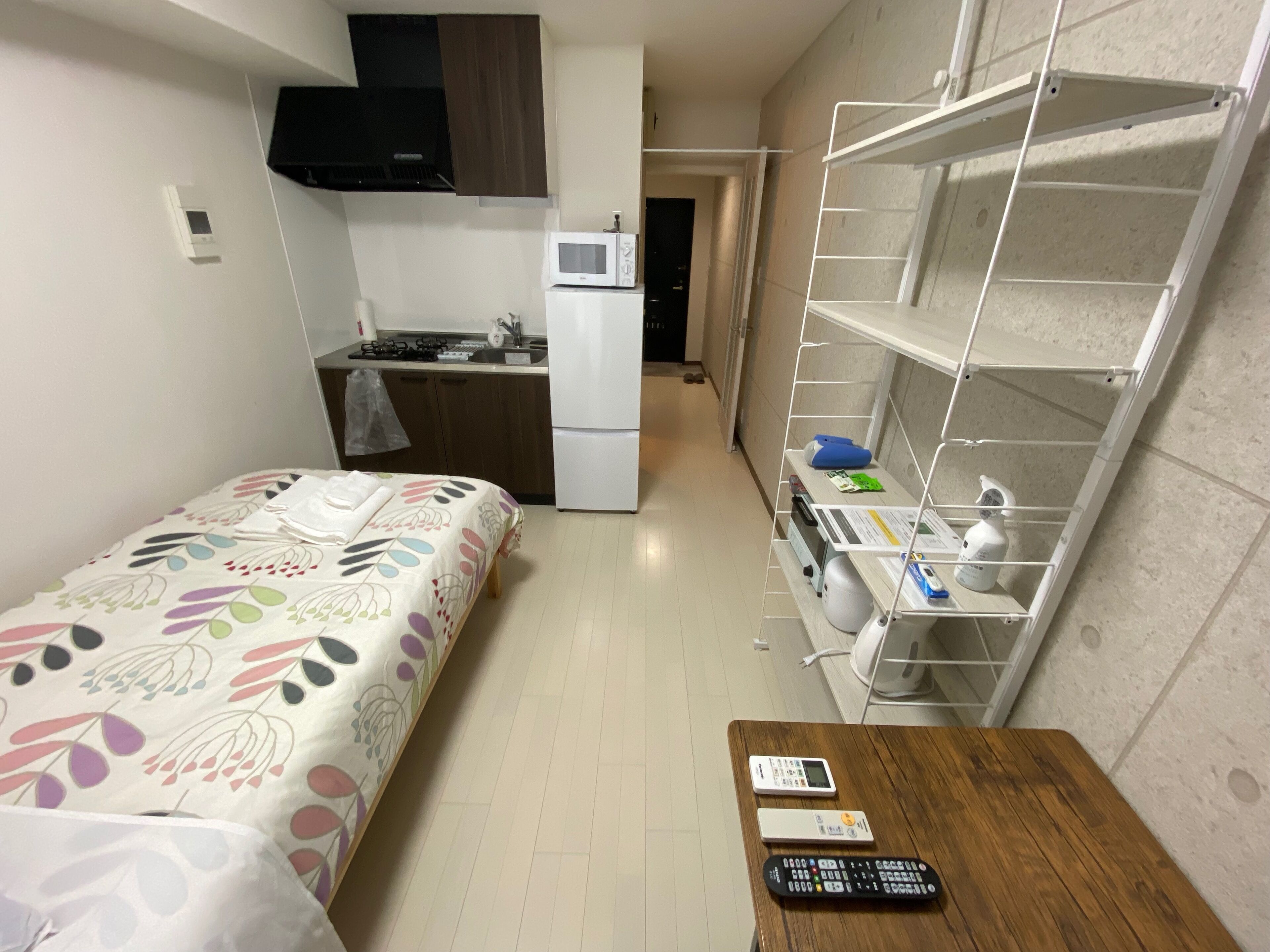 Cocina compacta con microondas, estufa y refrigerador. Hay una cama con cubrecama floral y una mesita con un control remoto.