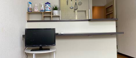 A compact kitchen with a built-in stove, a small table with a television, and a shelf with cleaning supplies.