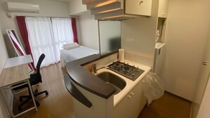 A compact kitchen with a gas stove, a sink, and a small countertop. There is a bed with white bedding and red curtains in the background. - White Terrace Sakurashinmachi (Tokyo)