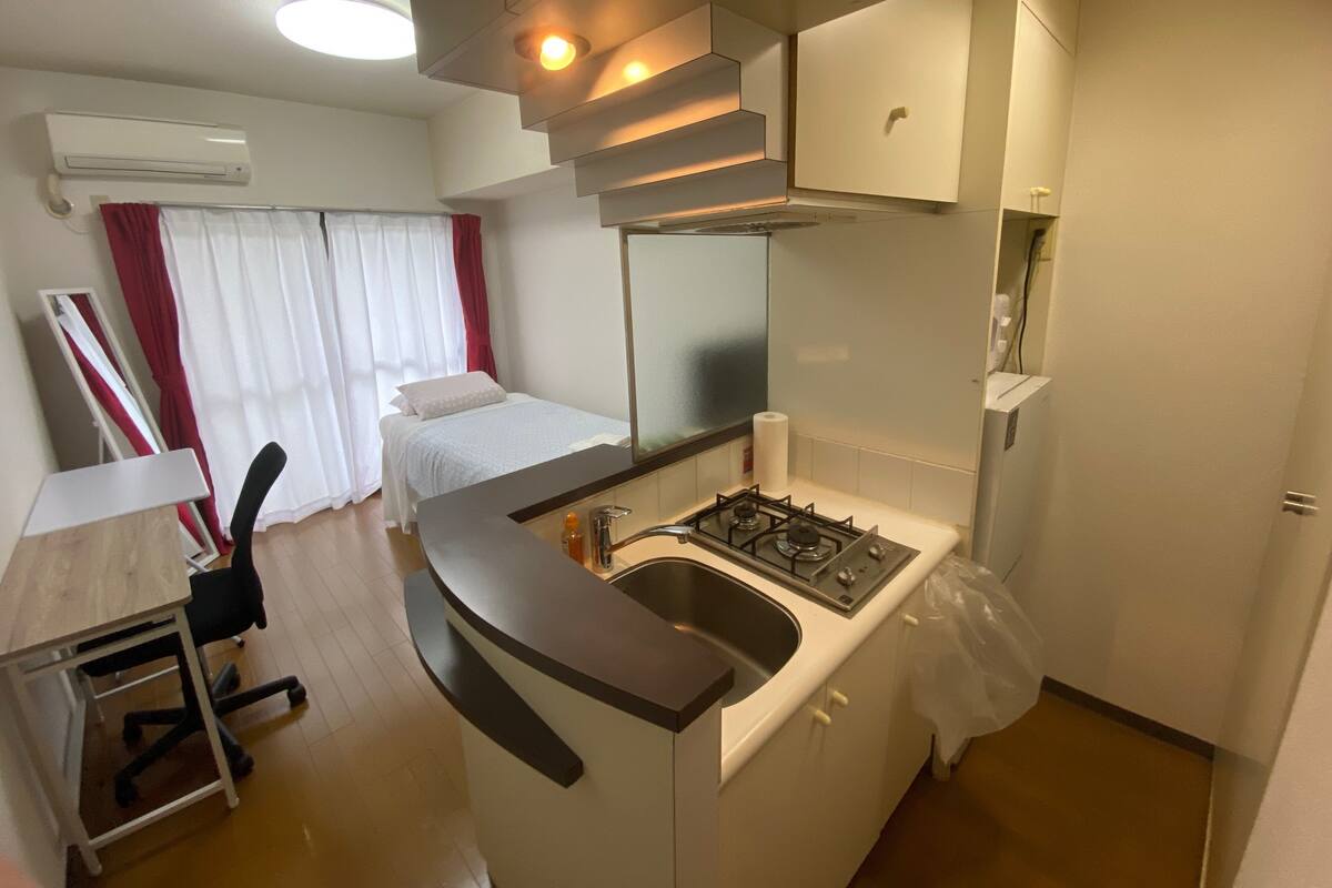 A compact kitchen with a gas stove, a sink, and a small countertop. There is a bed with white bedding and red curtains in the background.