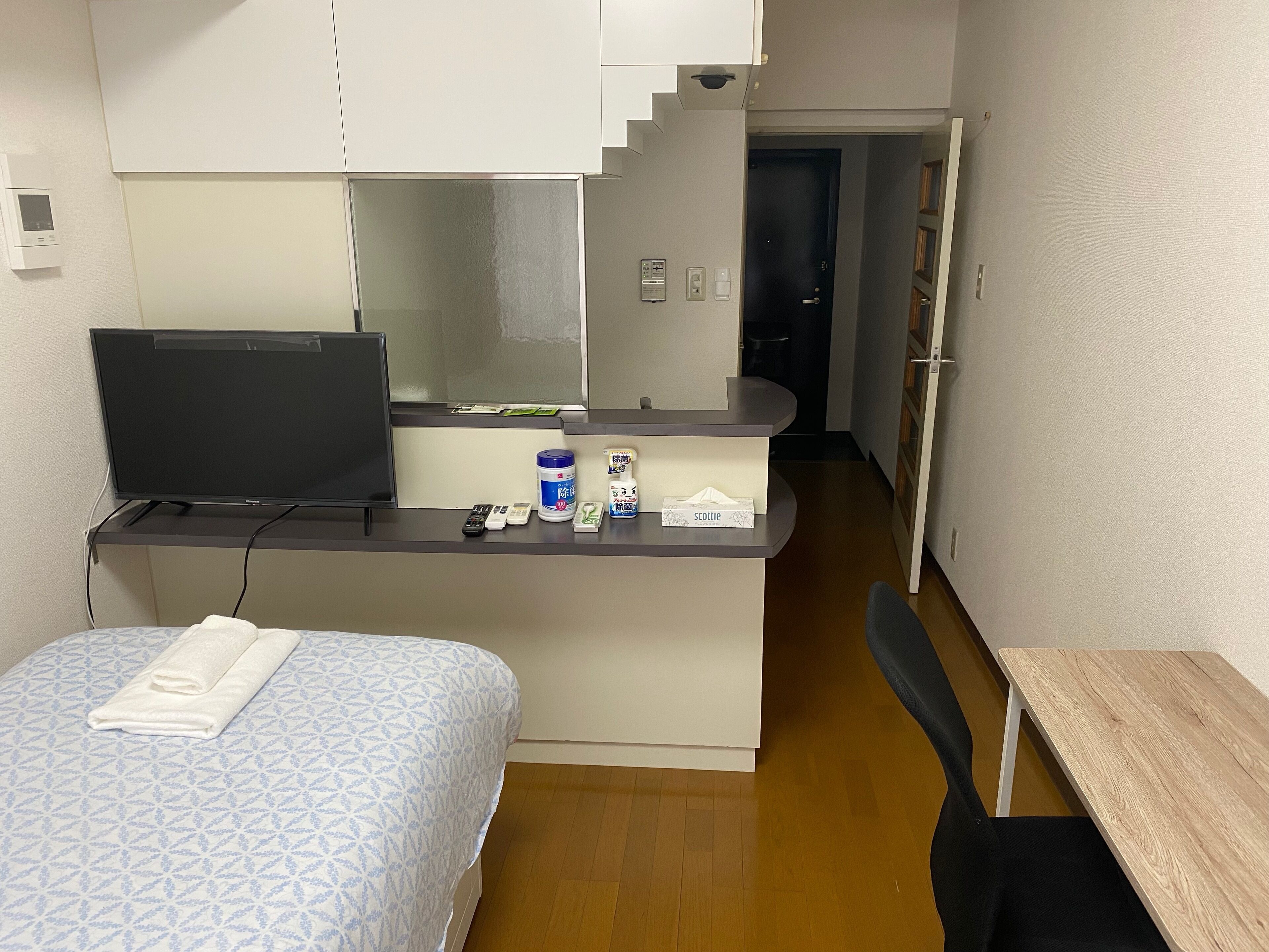 Una habitación de hotel compacta con cama, un escritorio con computadora, una kitchenette pequeña y un baño visible a través de una puerta abierta.