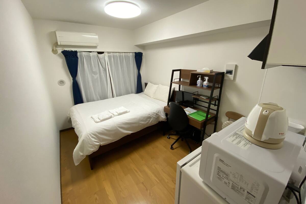 A compact hotel room with a single bed, a desk with a chair, a small kitchenette, and a ceiling-mounted air conditioner.