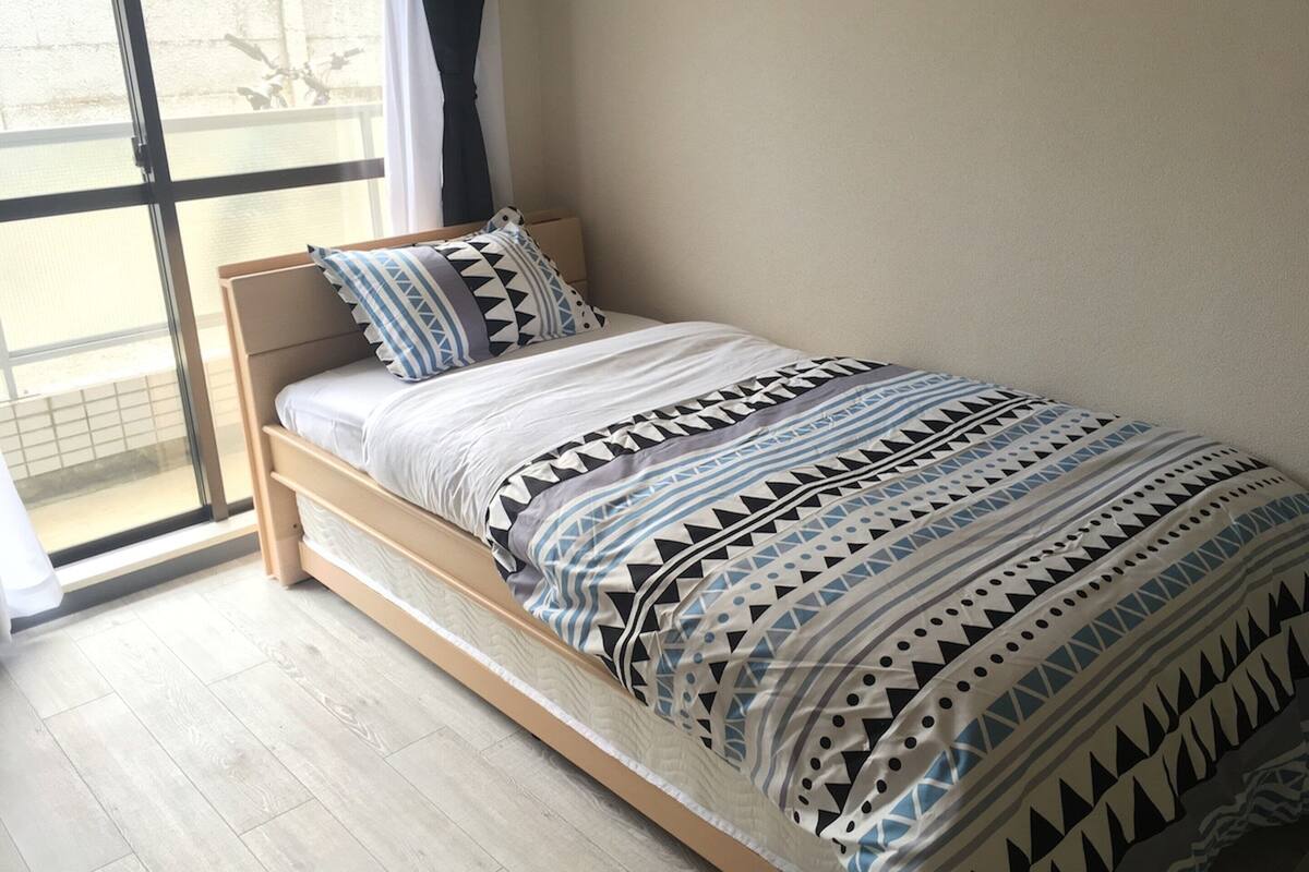 A single bed with a geometric patterned bedspread, a wooden headboard, and a window with a view of the outside.