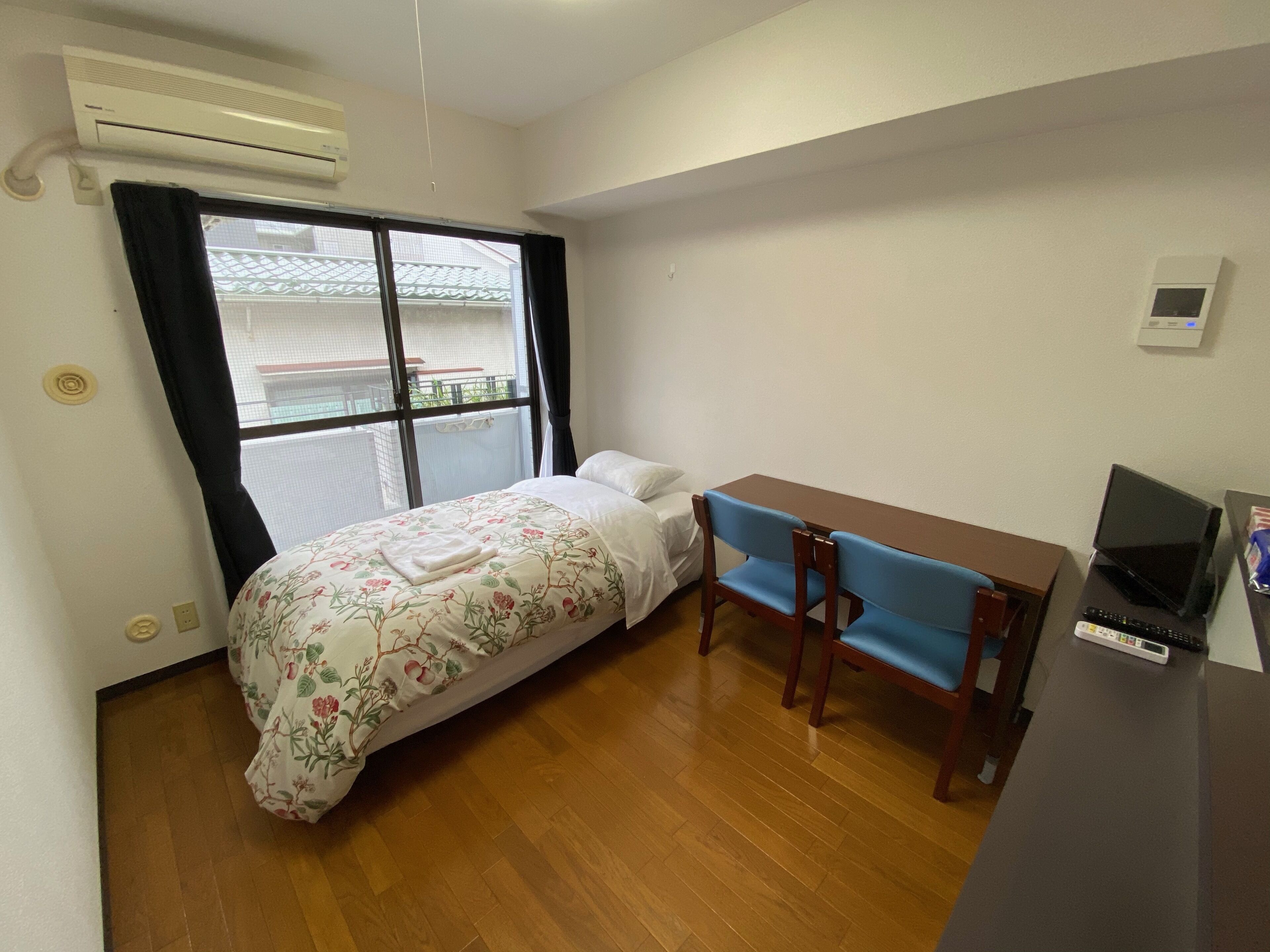 Une petite pièce propre, avec un lit, un bureau et une chaise. Il y a une fenêtre avec des rideaux, un climatiseur et un thermostat fixé au mur.