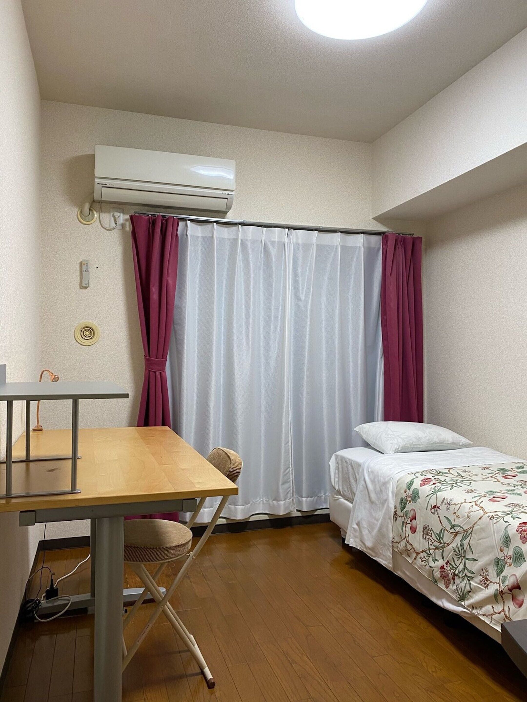 A compact room with a wooden table, a bed with a floral bedspread, a window with white curtains, and an air conditioning unit on the wall.