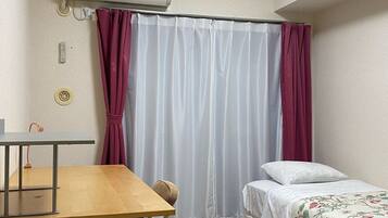 A compact room with a wooden table, a bed with a floral bedspread, a window with white curtains, and an air conditioning unit on the wall.