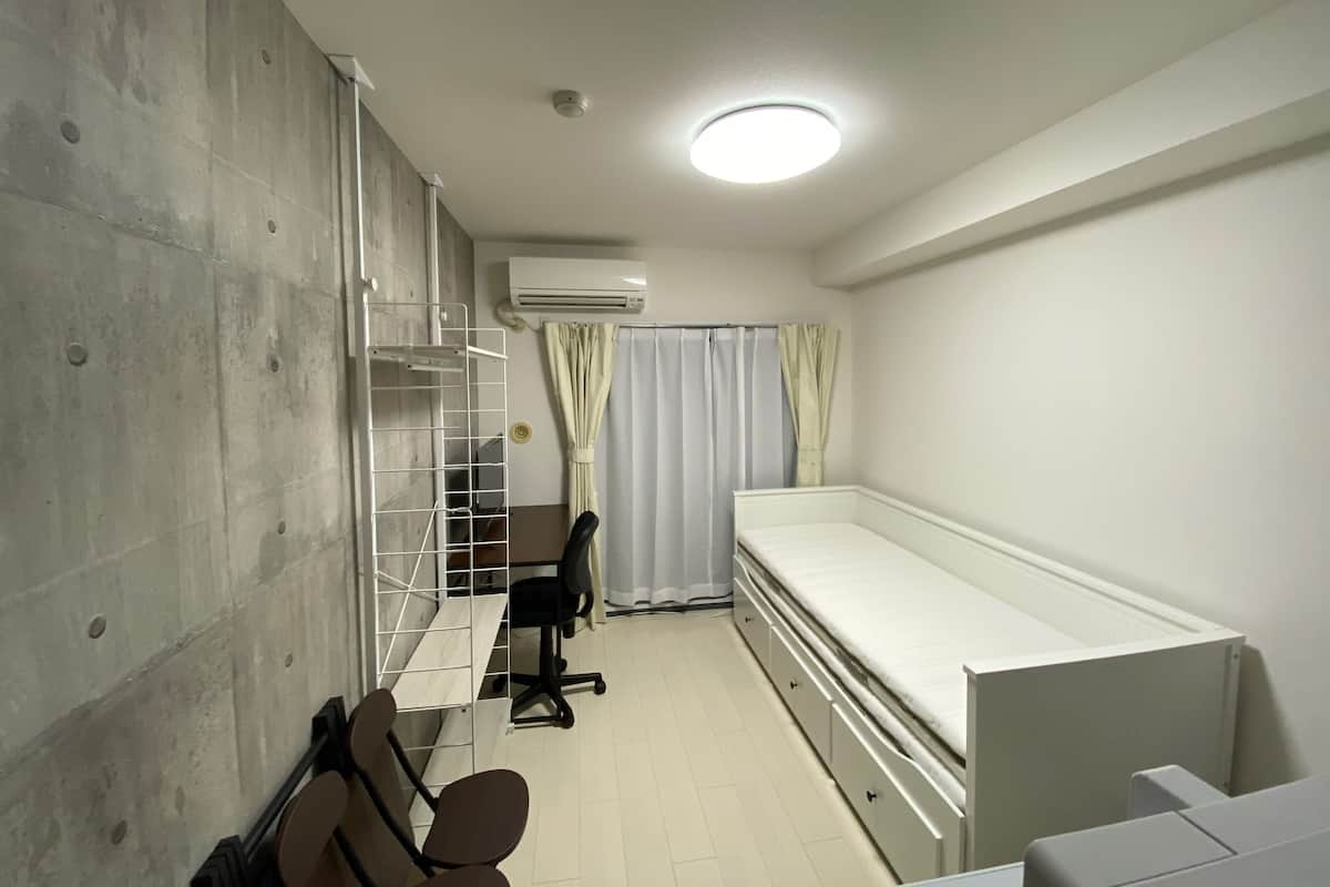 A clinical room with a single bed, a desk with a chair, a shelf unit, and a curtain.