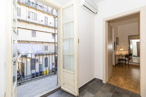 A room with a large window, a door leading to a balcony, a small table with chairs, and an air conditioning unit.