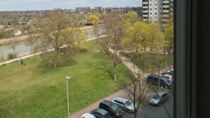 Vista da janela sobre um parque de estacionamento com vários carros, uma área ajardinada e edifícios ao longe.