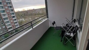 Uma varanda com carpete verde, uma mesa com cadeiras e vista para um edifício na rua.