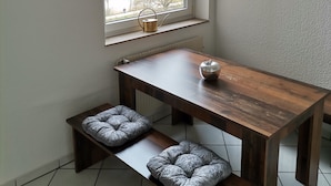 Quarto pequeno e moderno com mesa e banco de madeira, janela com vista para uma cidade e relógio de parede.