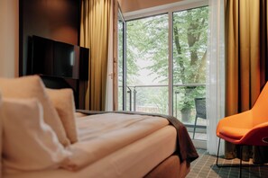 A hotel room with a bed, a television, a balcony with a chair, and a view of trees. - VIA PLAZA Meppen by Hackmann (Meppen)
