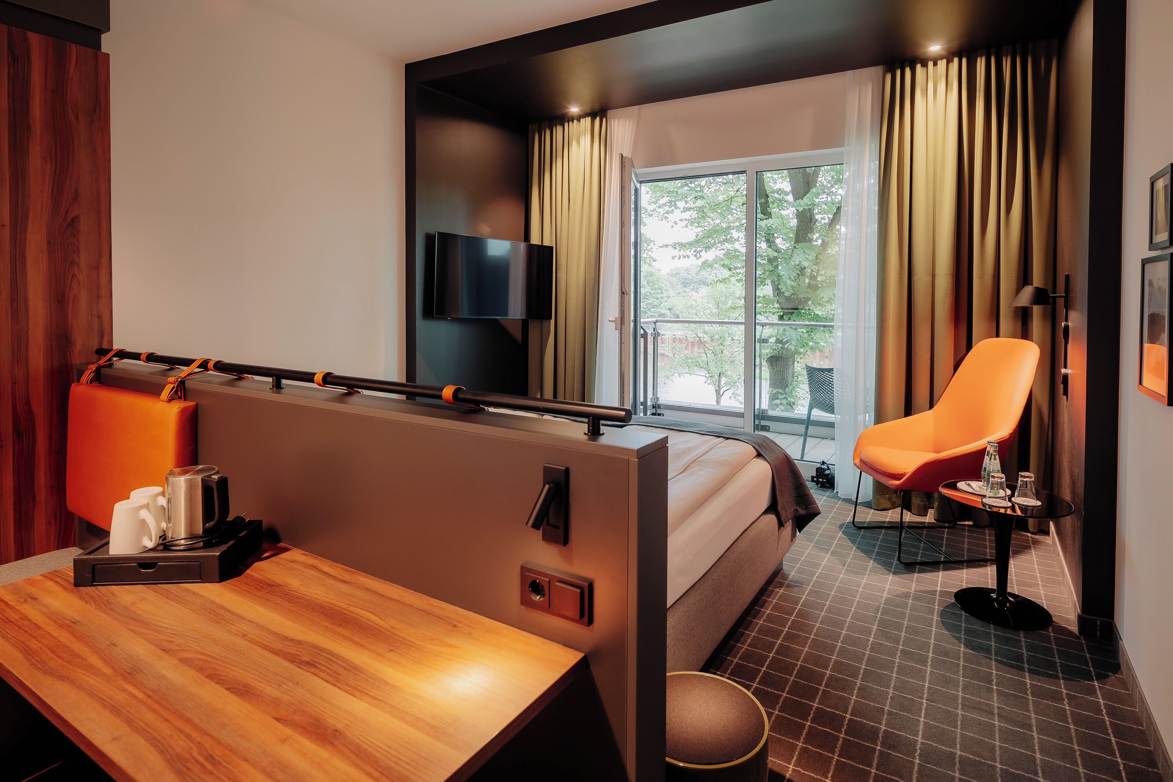 A modern hotel room with a wooden desk, a bed, a television, and a balcony with curtains.