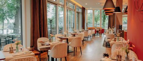 A spacious restaurant with high ceilings and large windows, offering a view of the outdoors. Tables are neatly arranged with chairs, and the interior features modern pendant lighting.