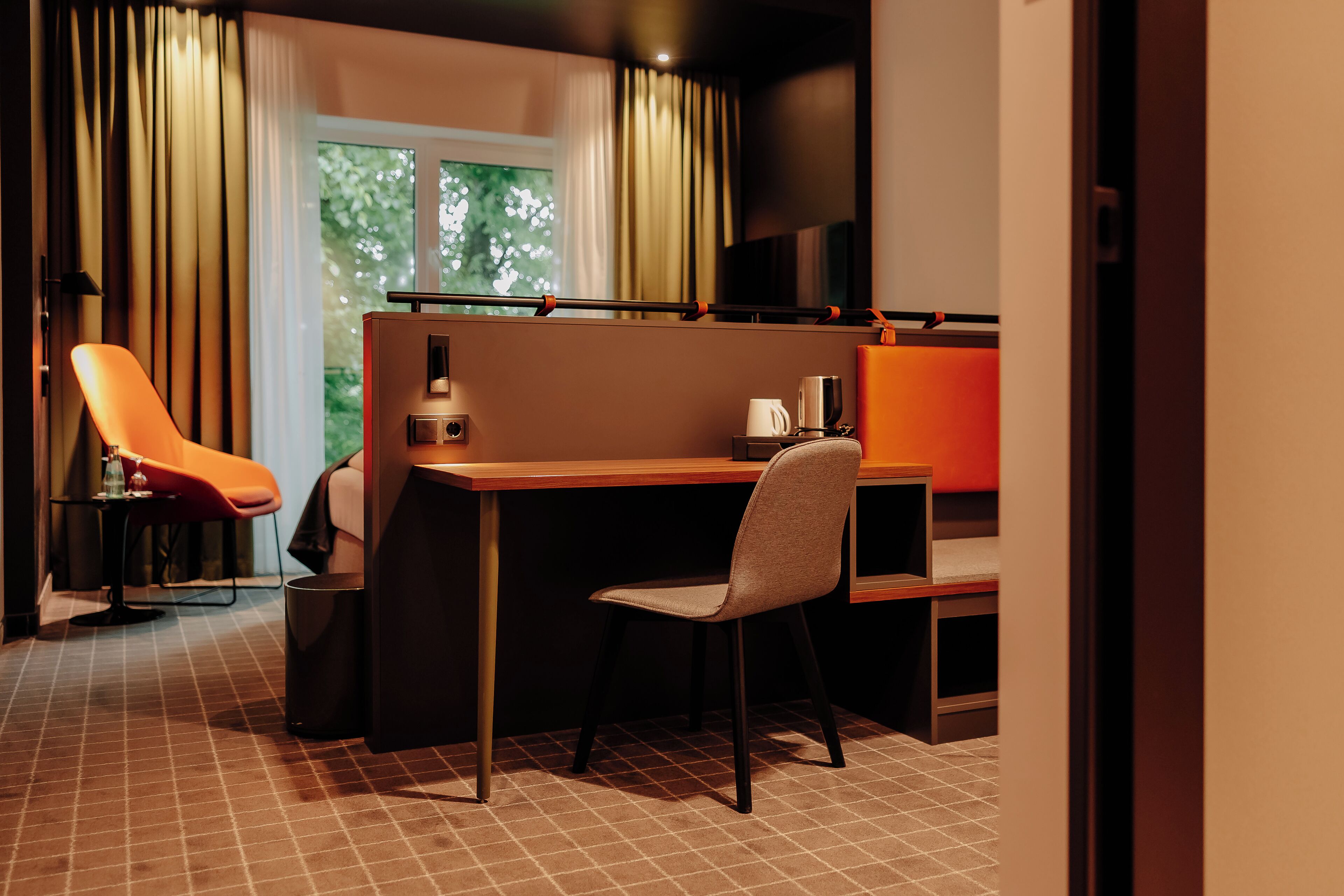 A modern hotel room with a desk, two chairs, a TV, and a window with curtains.