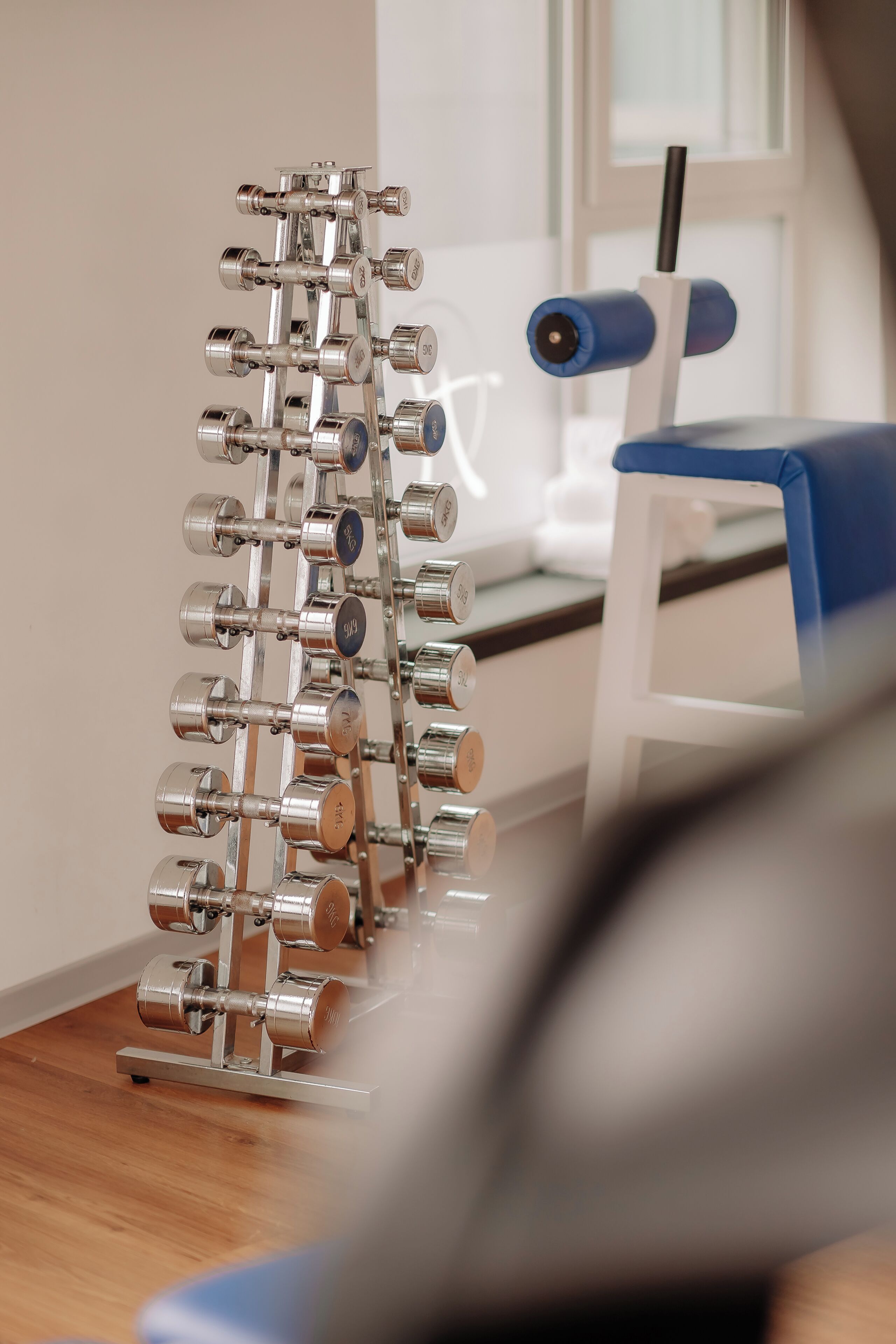 A metal dumbbell rack with weights, a blue chair, and a white exercise machine in a room with wooden flooring.