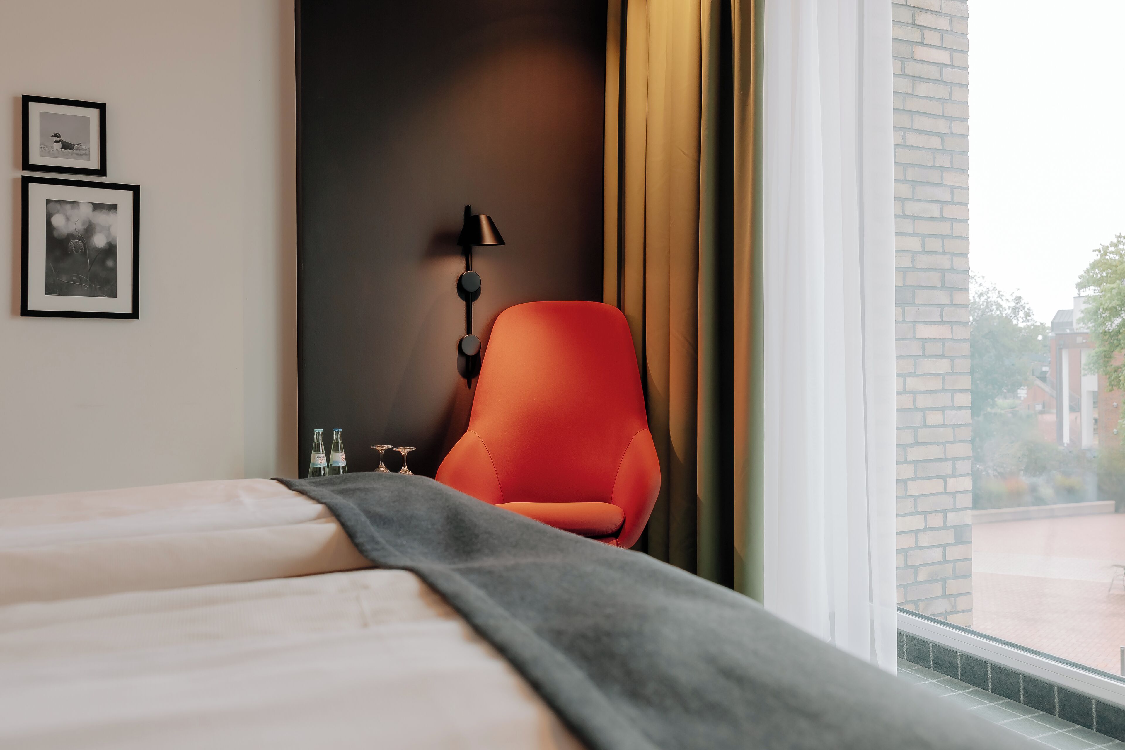 Hotellihuone, jossa on sänky, punainen tuoli, lamppu ja ikkuna, jossa on verhot.
