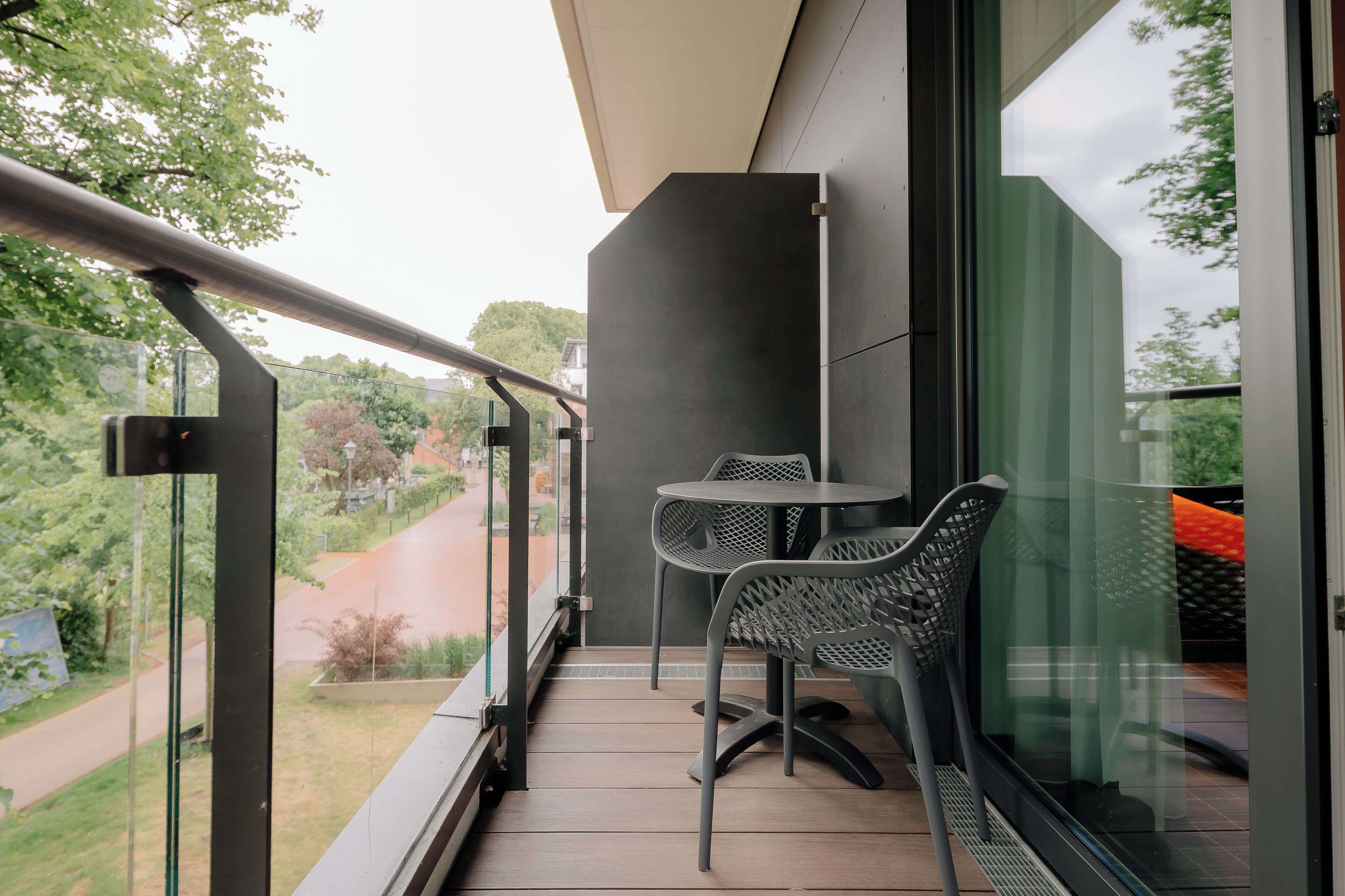 A balcony with a table and chairs, a glass door, and a view of a road and greenery.