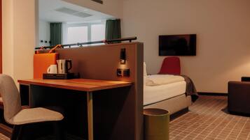 Una habitación de hotel con cama, escritorio, silla, televisor y un cubo de basura.