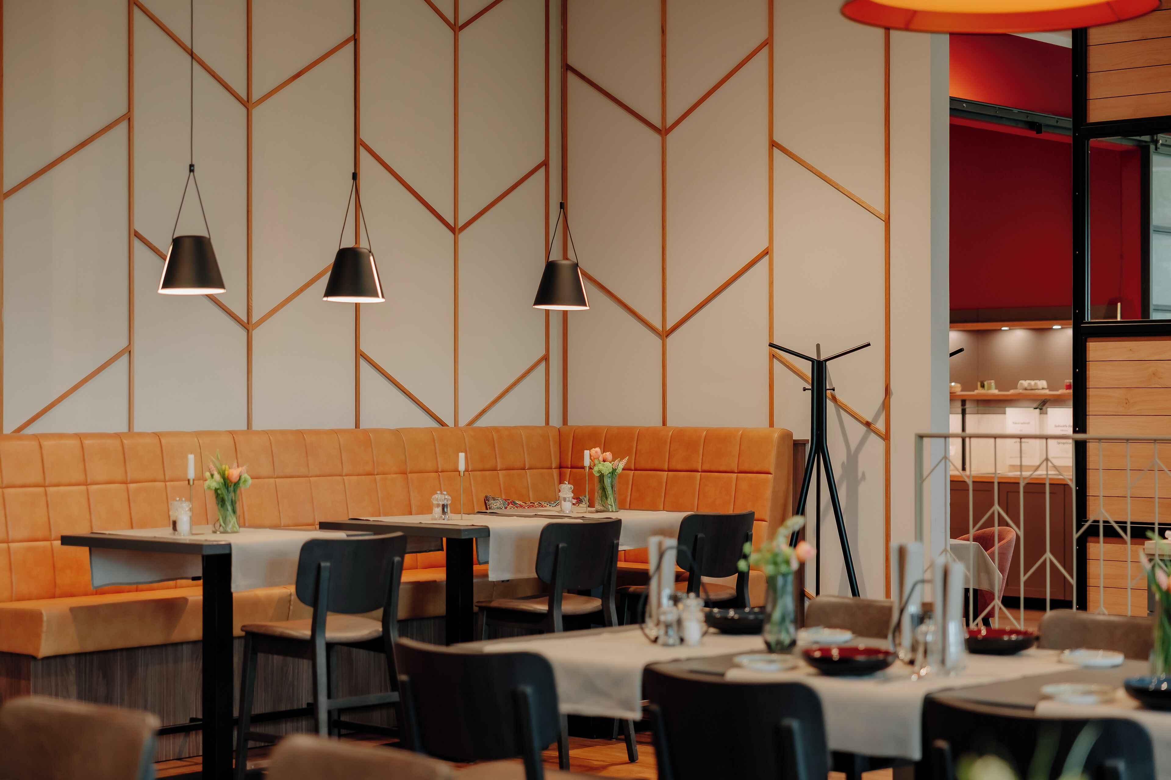 A restaurant with a modern interior, featuring geometric wall patterns, orange seating, and black pendant lighting.