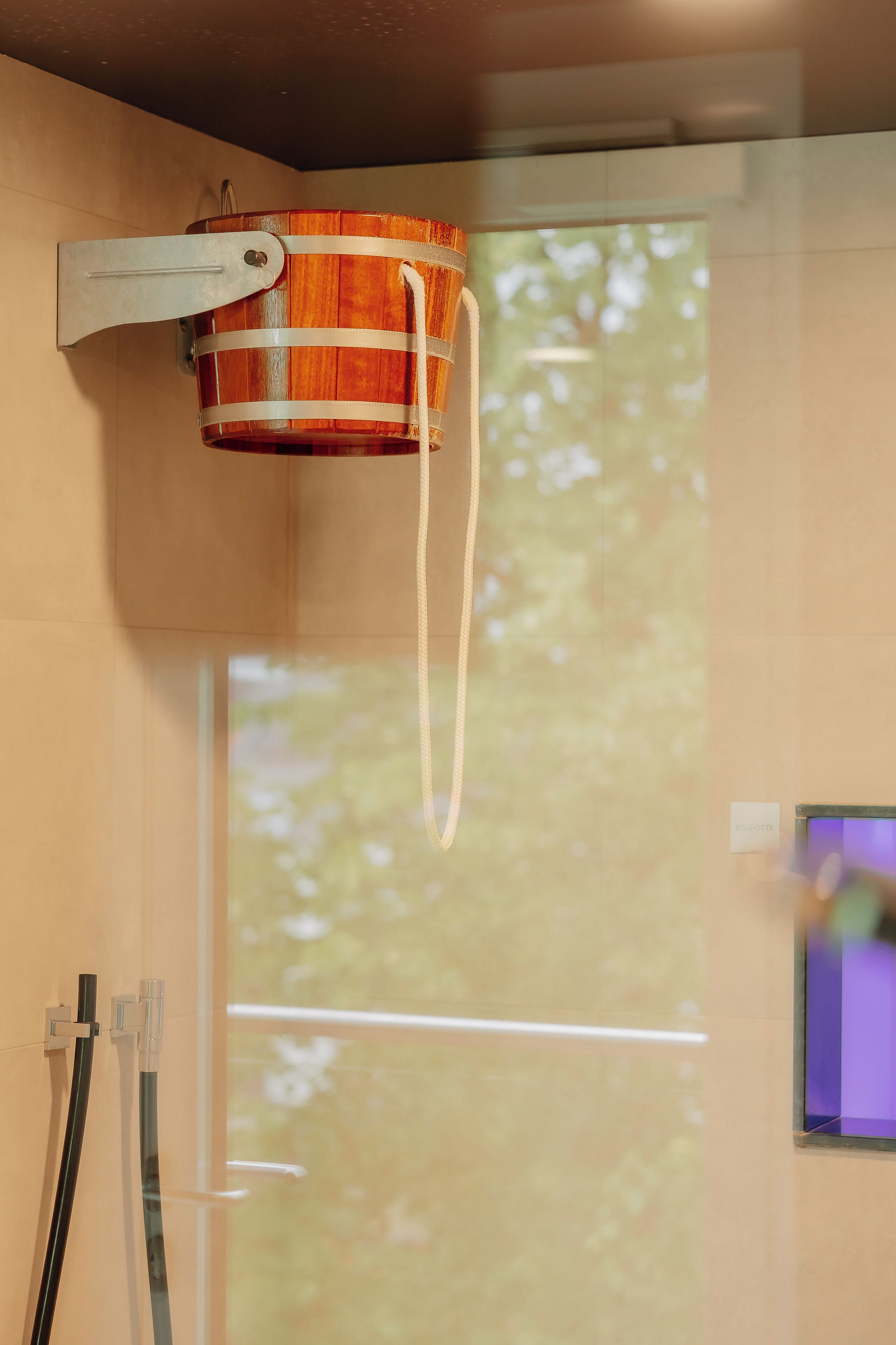 A wooden bucket mounted on a wall with a handle, a window with a view of trees, and a small screen displaying an image.