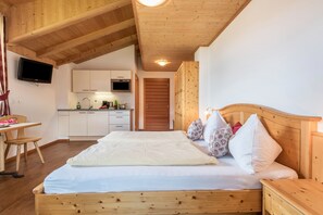 Un dormitorio con una cama de madera, una mesita de noche, una pequeña alacena junto a la cama, un televisor montado en la pared, una zona de cocina con armarios y un microondas, y techo de madera.
